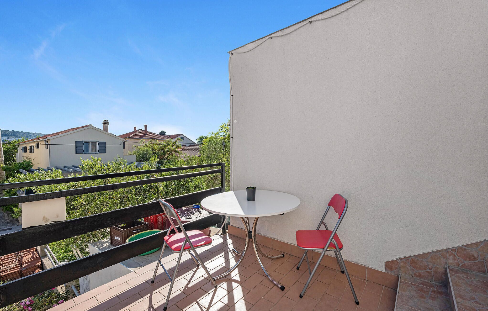 Un balcón con una mesa blanca y dos sillas rojas, con vistas a una zona residencial con casas y vegetación.