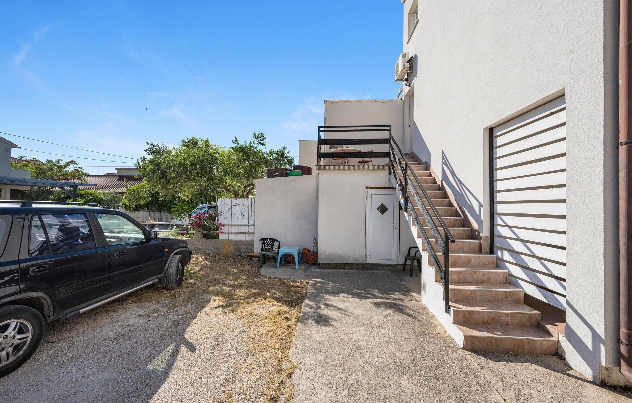 A residential area with a black car parked on a gravel driveway, a staircase leading to a garage, and a small outdoor seating area with a blue chair and a table.