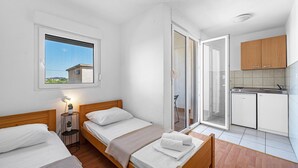 Ein kleiner, heller Raum mit zwei Betten, einem Fenster, einer Küchenzeile und einer Glasschiebetür zu einem Außenbereich.