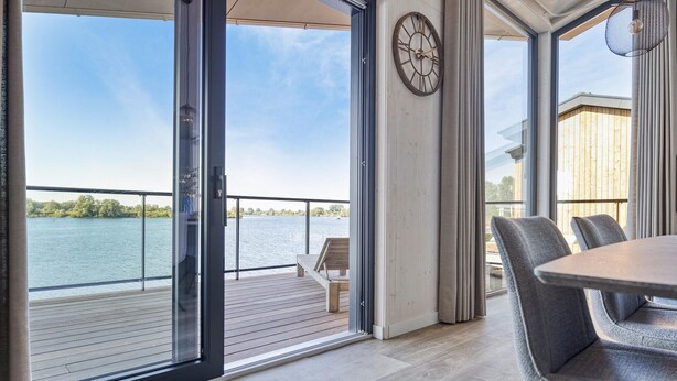 Une chambre moderne dotée d’une grande porte-fenêtre coulissante qui donne sur une terrasse offrant une vue sur un lac. On y trouve une horloge au mur et une table avec des chaises.