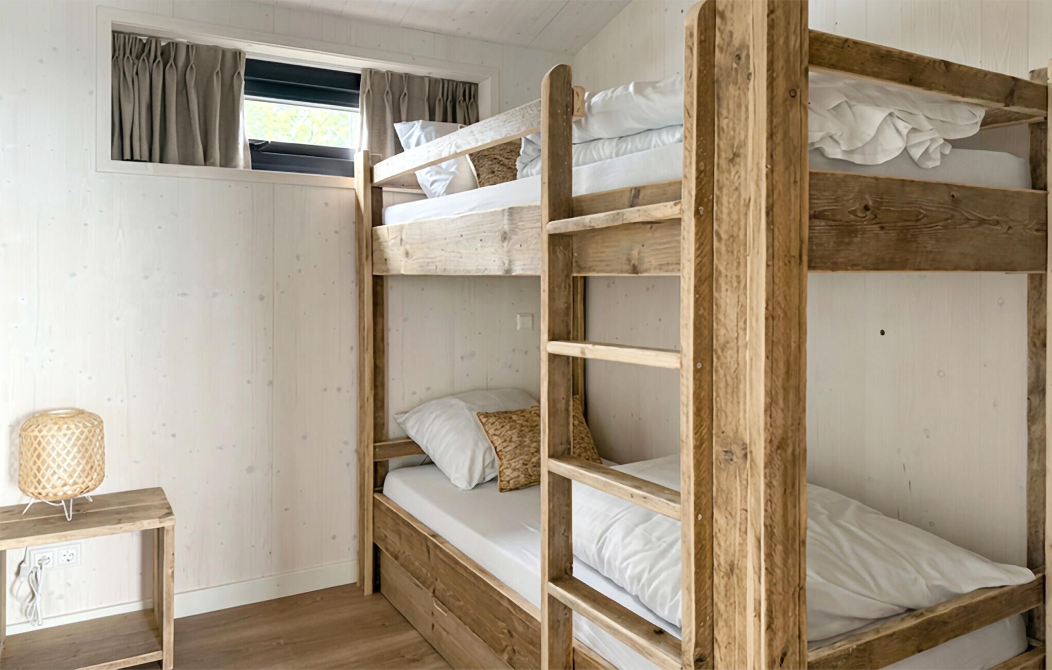A bunk bed room with a window, a small table with a lamp, and a plain wall.
