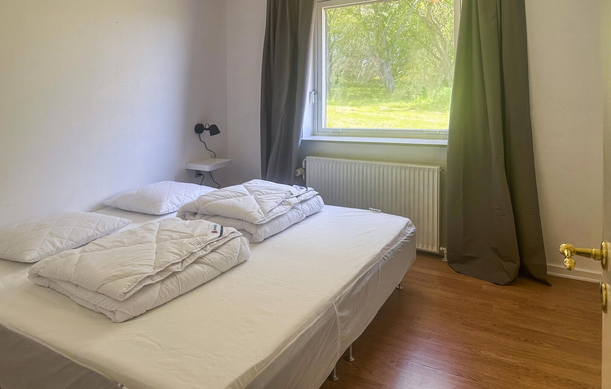 En enkel seng med hvide sengetøj og sengegavl, et natbord med lampe, et vindue med gardiner og en radiator.