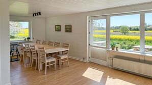Un espace repas avec une table et des chaises en bois, de grandes fenêtres donnant sur un champ, et un plafond doté d’un éclairage encastré.