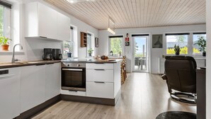 A modern kitchen with white cabinetry, a built-in oven, and a coffee maker. There is a dining area with a table and chairs, and a view of the outdoors through large windows. - 3 bedroom cozy home in Højslev (Højslev)