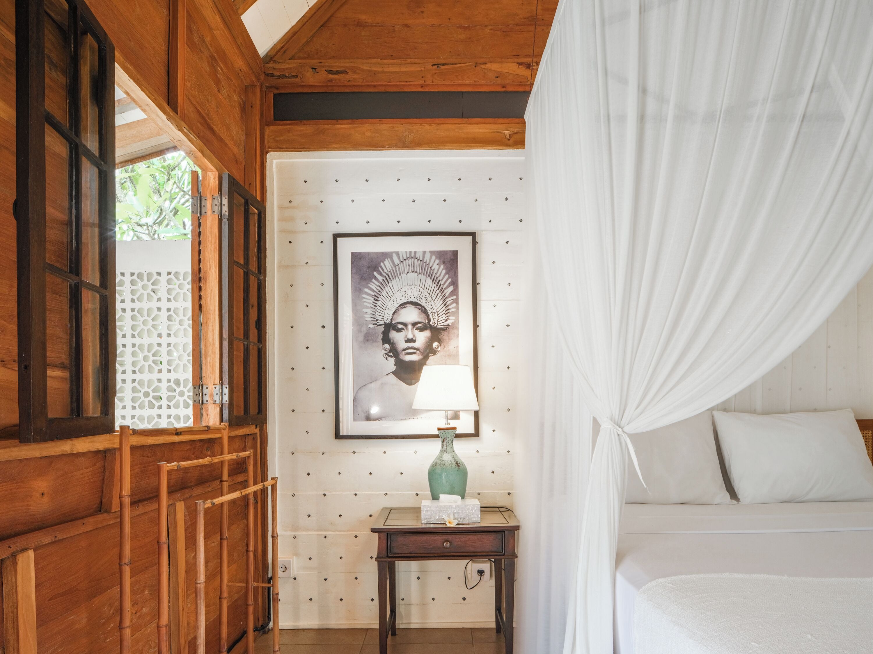 Een houten kamer met een bed, nachtkastje, een ingelijste foto en een raam met uitzicht op groen.