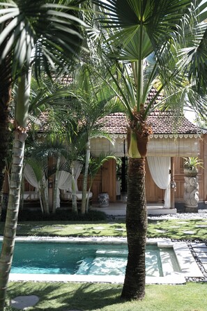 A poolside area with palm trees, a tiled pool, and a building with a tiled roof and wooden doors. - Nido Boutique Cottage (Canggu)