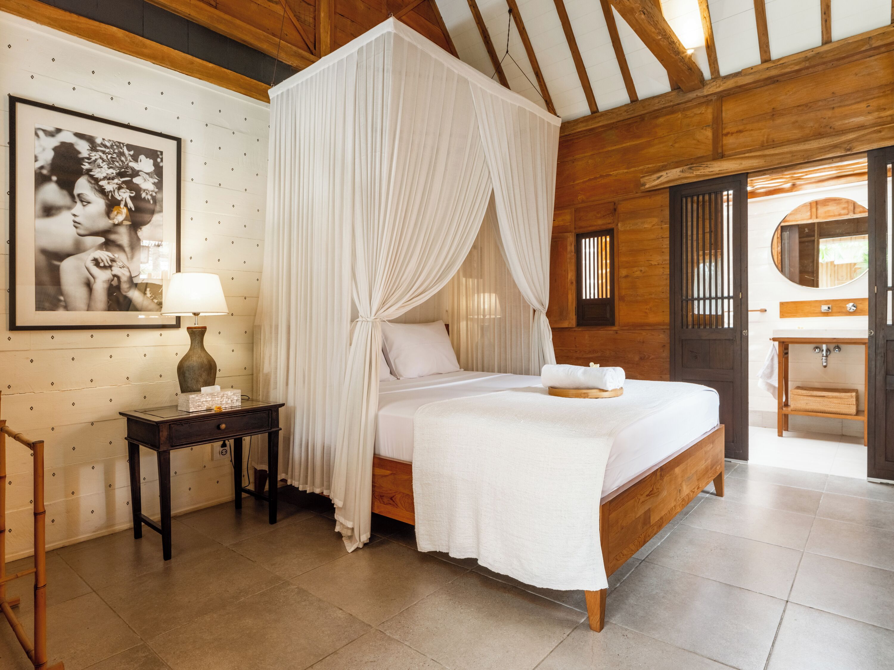 A bedroom with a four-poster canopy bed, a bedside table with a lamp, a framed picture on the wall, and a wooden door leading to a bathroom.