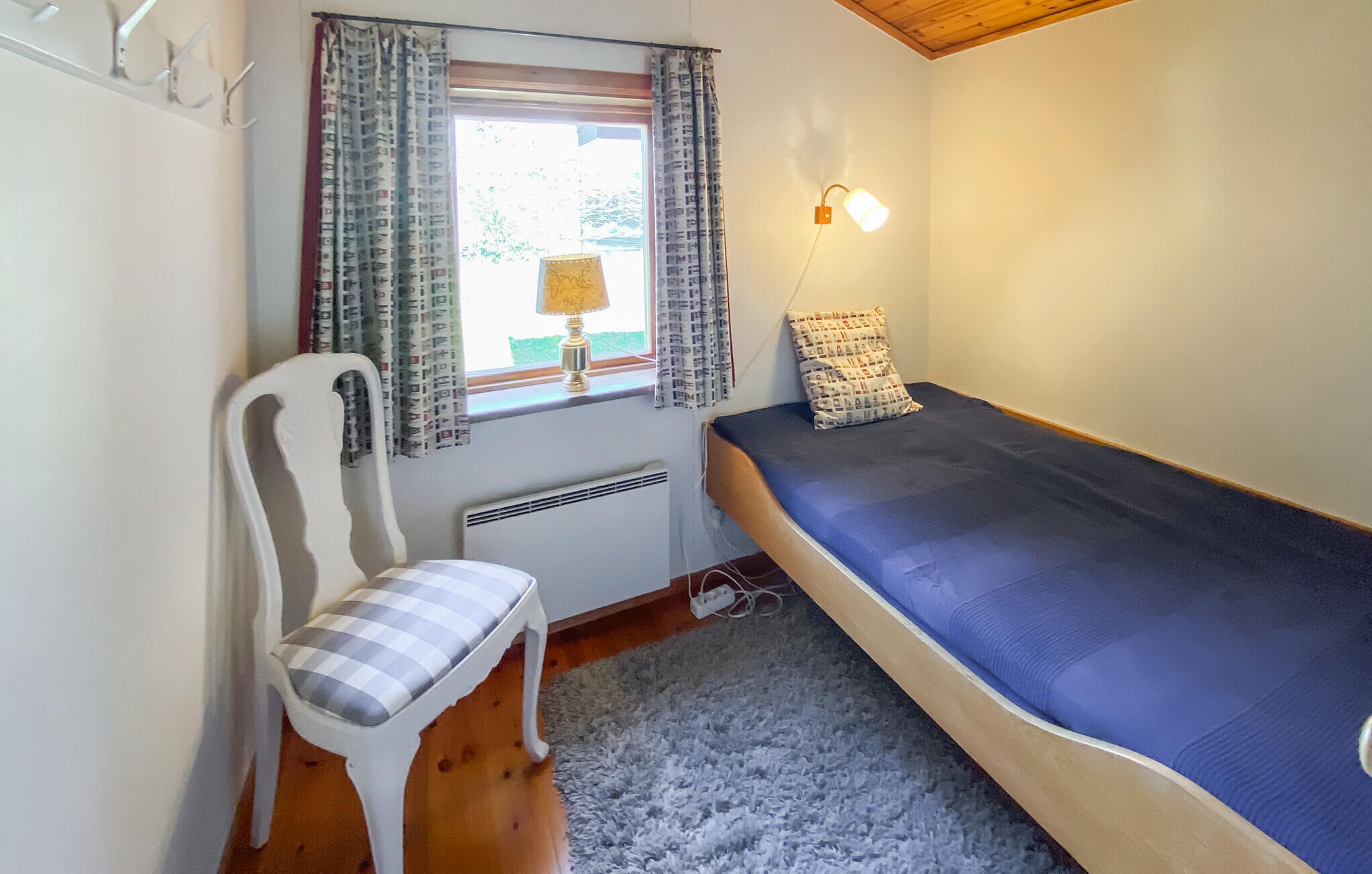 Una stanza piccola con un letto singolo, una sedia, una finestra con tende, una lampada a muro e un termosifone.