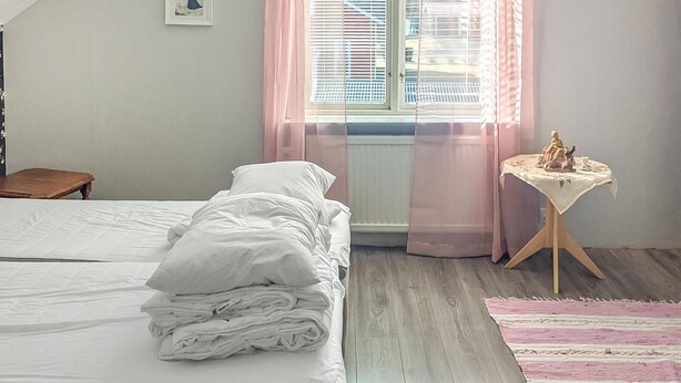 A neatly made bed with white linens, a small wooden nightstand with a statue, and a pink-striped rug on a wooden floor. - Cozy home in Bastuträsk with WiFi (Bastuträsk)