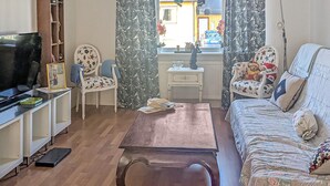 A living room with a wooden coffee table, a sofa with floral upholstery, a television on a stand, and a window with curtains. - Cozy home in Bastuträsk with WiFi (Bastuträsk)