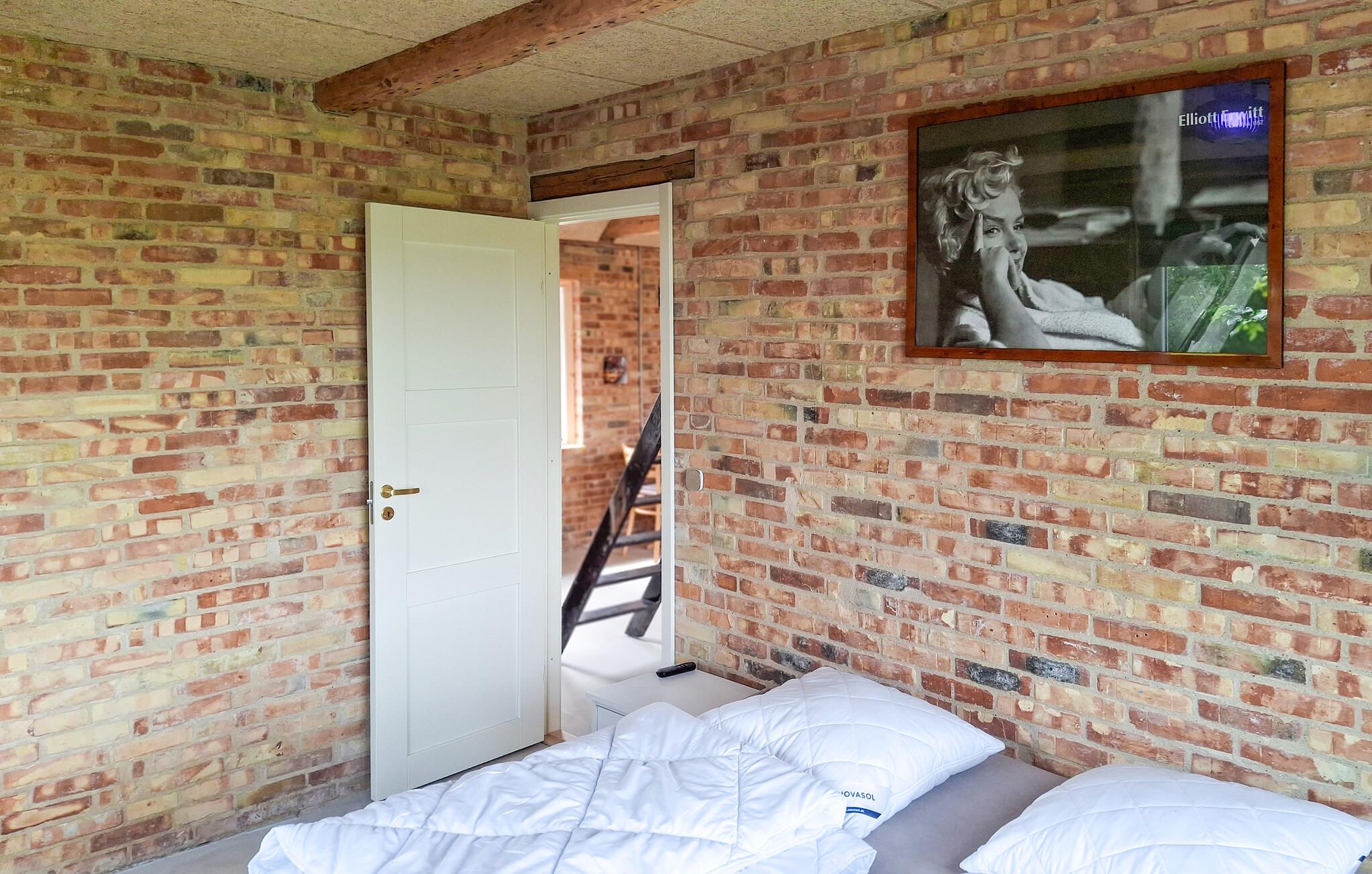 Una habitación con pared de ladrillo, una puerta blanca, una cama con ropa de cama blanca y un cuadro enmarcado en la pared.
