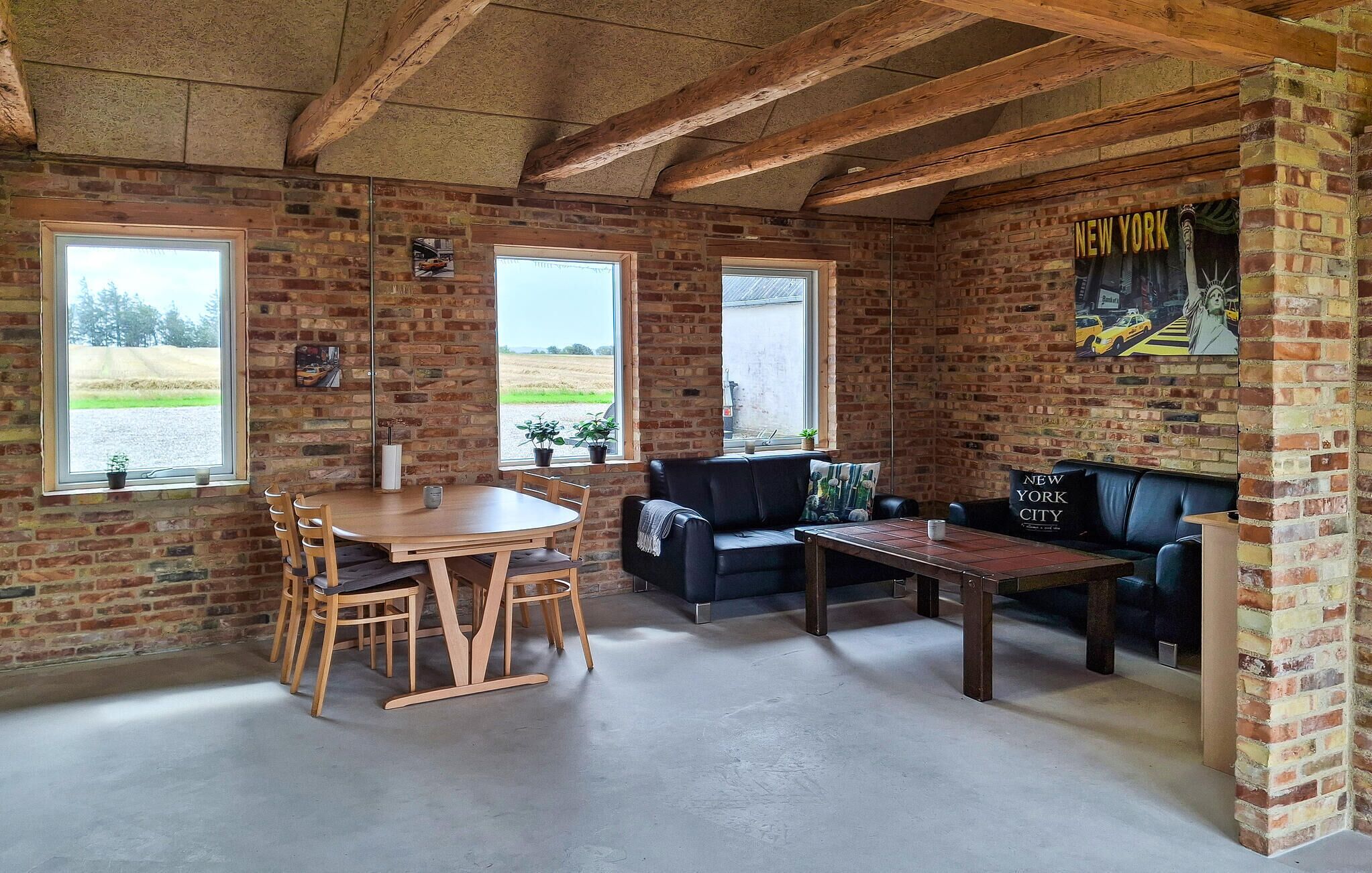 Un intérieur rustique avec des murs en briques apparentes, un plafond en bois, une table ronde en bois entourée de chaises, un canapé en cuir noir et une petite table basse en bois.