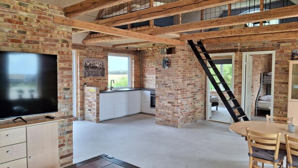 A spacious room with exposed wooden beams, a brick wall, a large flat-screen TV, a kitchen area, a dining table with chairs, and a staircase leading to an upper level. - Nice home in Erslev with WiFi (Erslev)