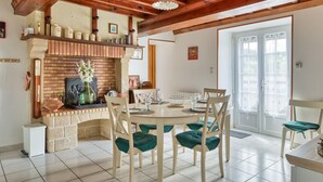 A dining area with a brick fireplace, wooden ceiling beams, and a large window. - Charmante maison de Campagne (Méautis)