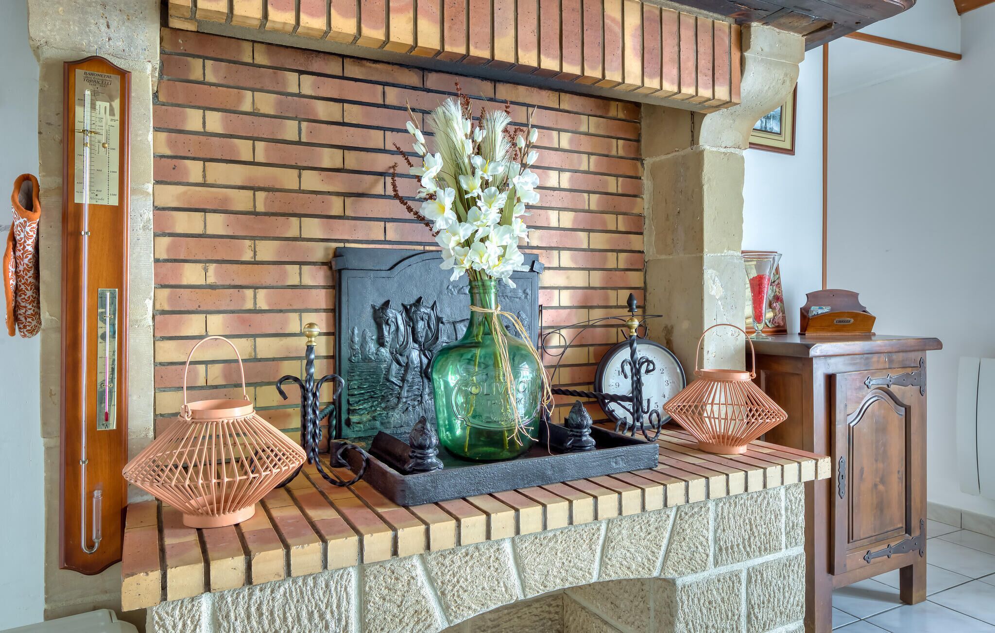 Una chimenea de ladrillo con una botella de vidrio verde y flores blancas sobre la repisa, un reloj de madera y un farol de cobre.