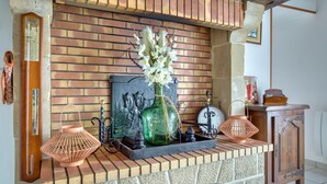 A brick fireplace with a green glass bottle and white flowers on the mantle, a wooden clock, and a copper lantern. - Charmante maison de Campagne (Méautis)