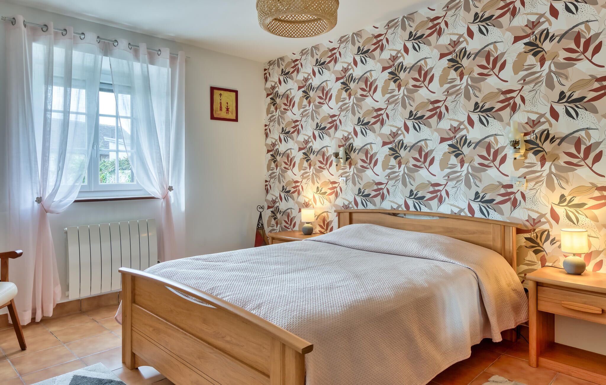 A bedroom with a wooden bed, a radiator, a window with sheer curtains, a wall with a leaf-pattern wallpaper, and bedside lamps.