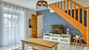 A modern living room with a wooden staircase, a TV on a white cabinet, and a dining table. - Charmante maison de Campagne (Méautis)