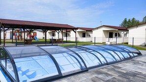A covered carport with a transparent roof, adjacent to a playground with a slide and swings, and a row of white buildings with red roofs. - Awesome home in Przewloka with swimming pool (Przewloka)