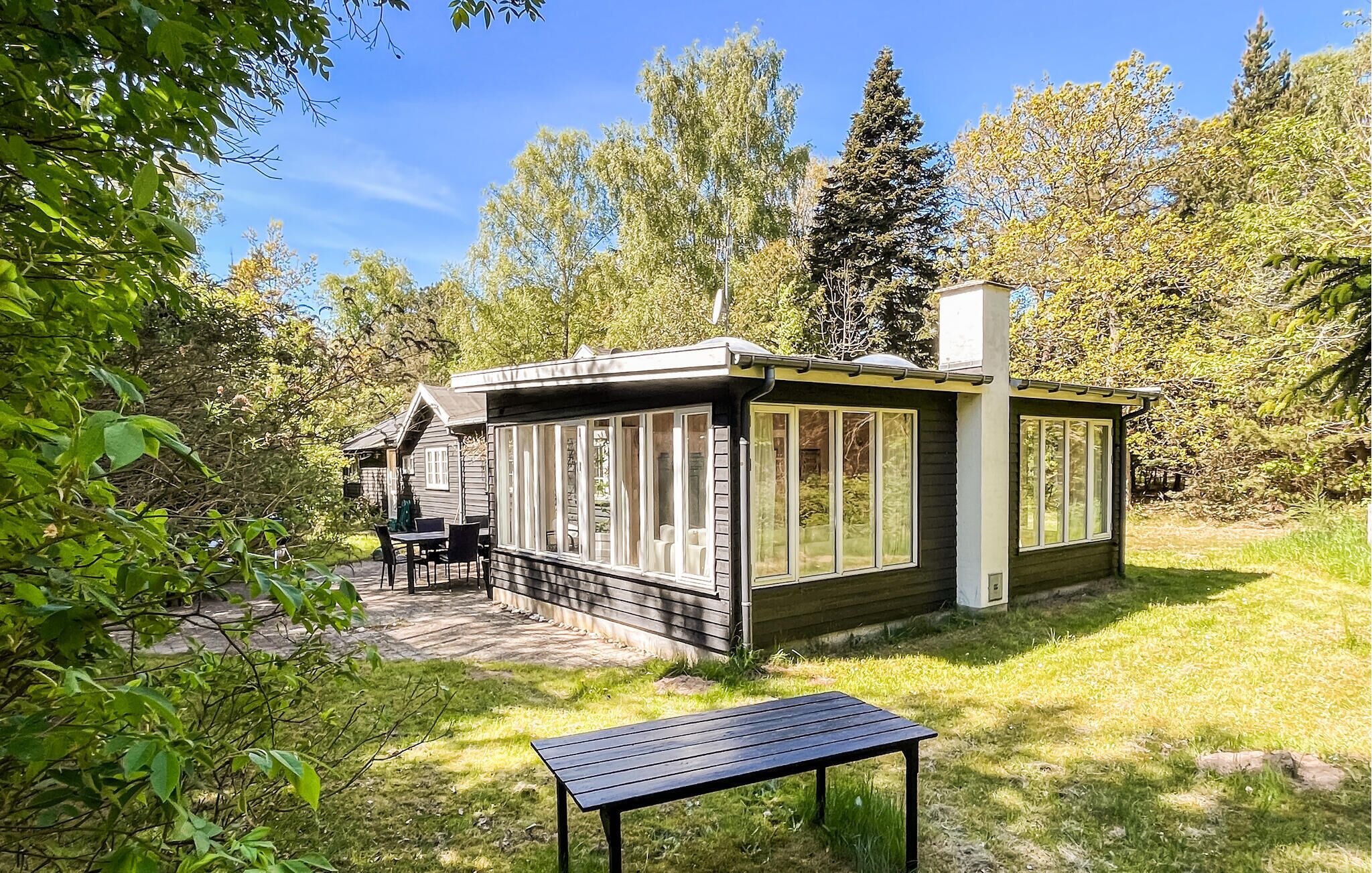 A small wooden cabin with large windows, a patio with a table and chairs, surrounded by trees.