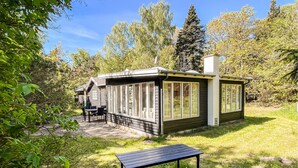 Una cabaña de madera pequeña con ventanas grandes, un patio con mesa y sillas, rodeada de árboles.