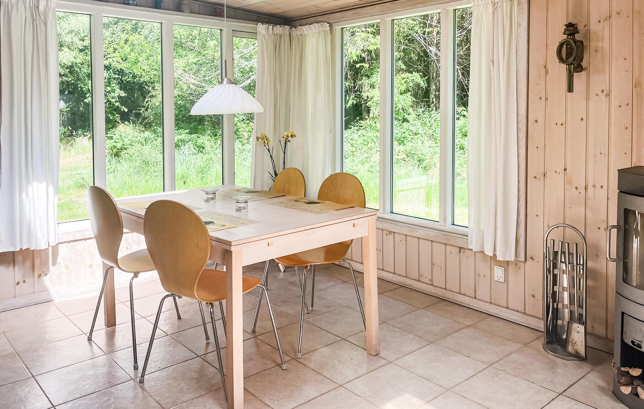 Un coin repas avec une table et des chaises en bois, de grandes fenêtres avec des rideaux et un poêle à bois.