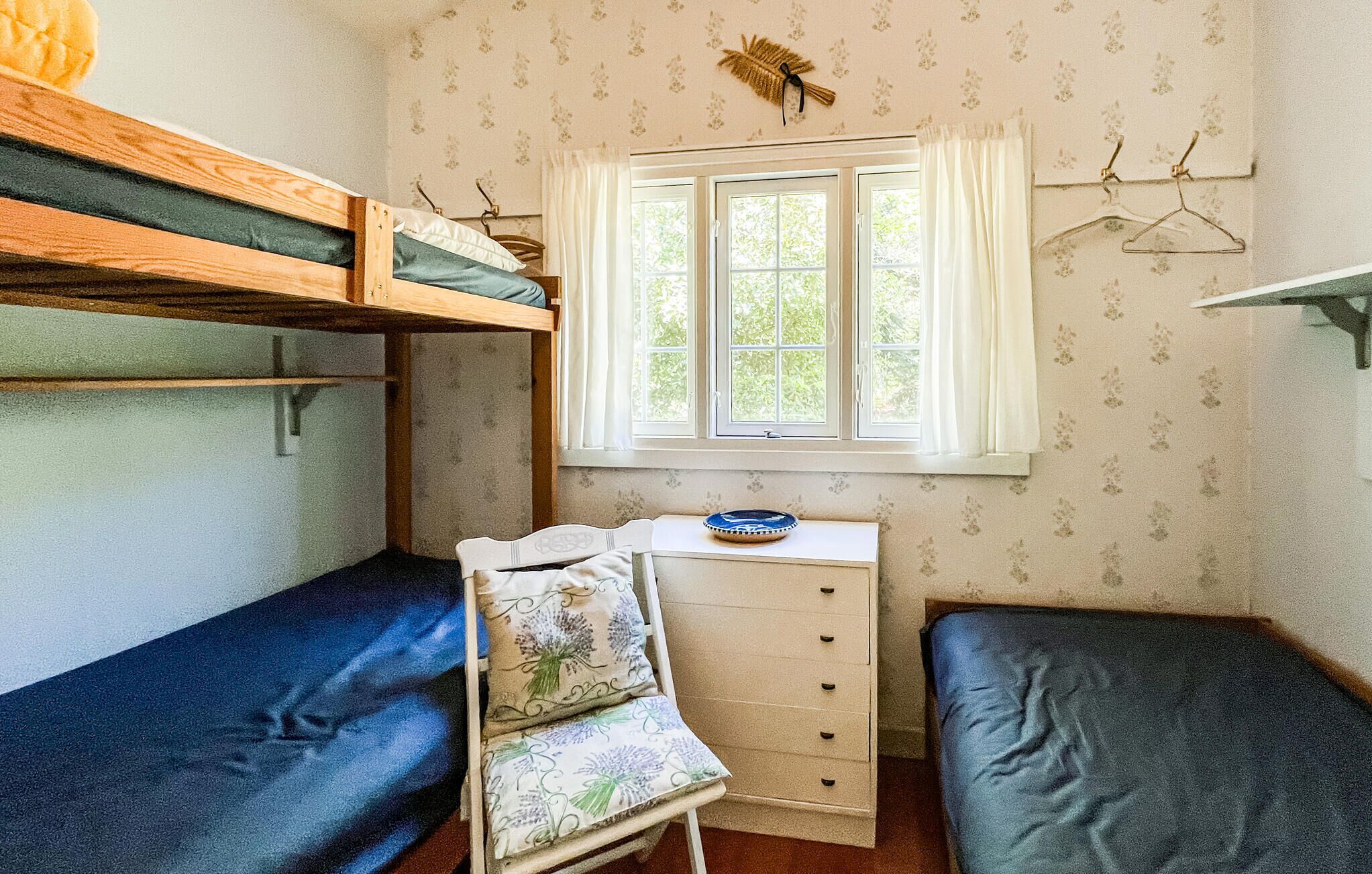 A dormitory room with bunk beds, a desk with drawers, a window with curtains, and a wall-mounted coat rack.