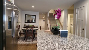A kitchen with a granite countertop, a refrigerator, a dining table with chairs, a potted plant, and a staircase leading to another room.