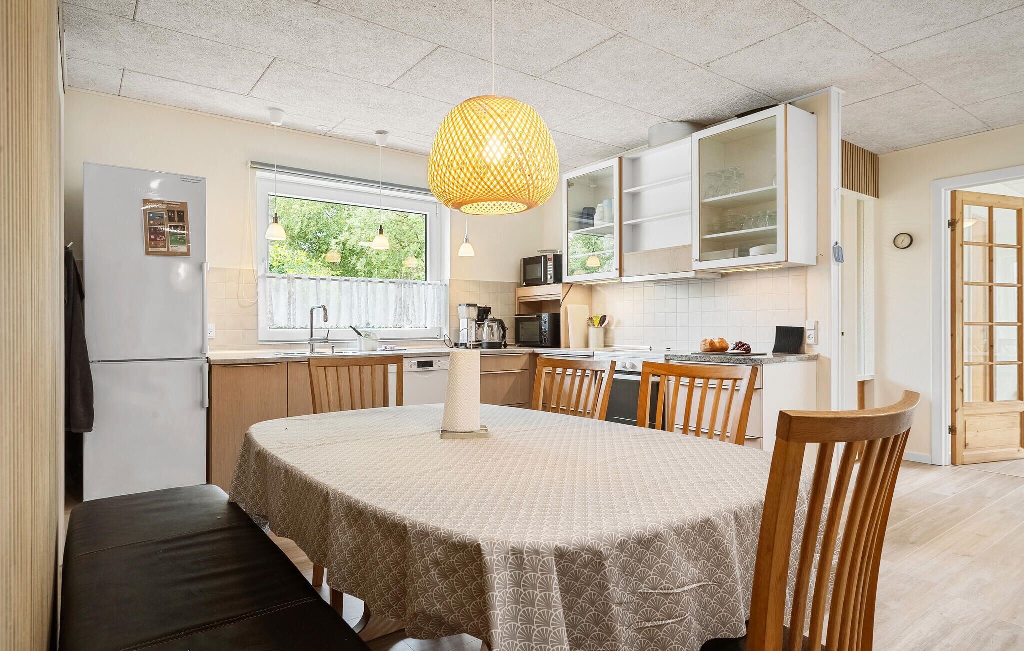 Een moderne keuken met witte kastjes, een eettafel met een tafelkleed met patroon en een hangende hanglamp.
