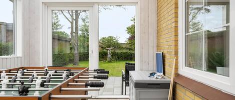 A room with a foosball table, a small table with items, and a window overlooking a garden.