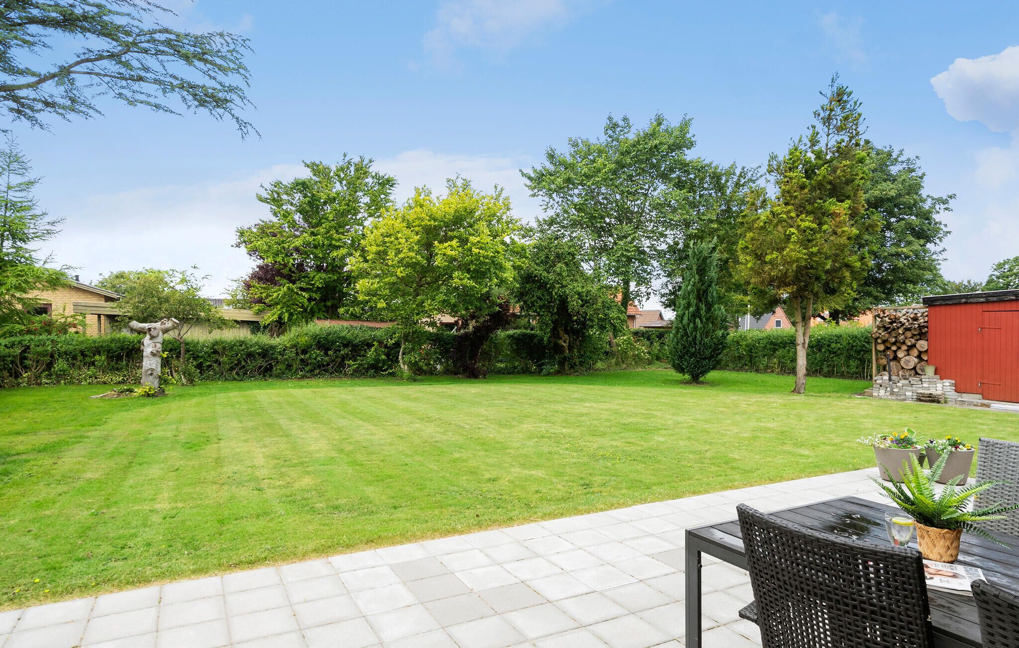 Een verzorgde tuin met gazon, een terras met tafel en stoelen, en een rood gebouw op de achtergrond.
