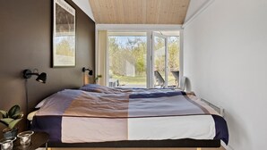 A bedroom with a bed, a nightstand, a lamp, a framed picture, and a view of the outdoors.