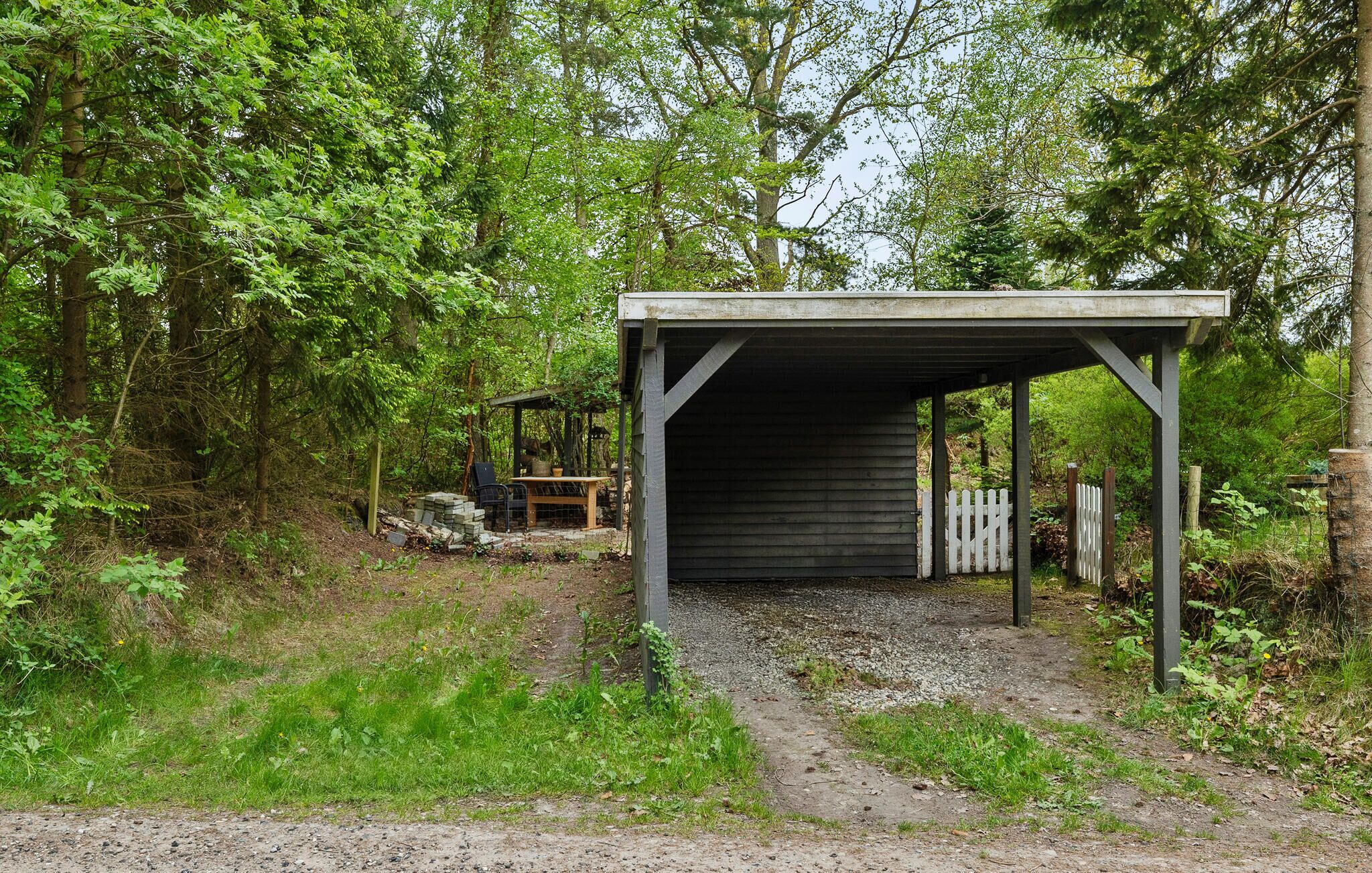 A covered outdoor area with a bench and a table, surrounded by trees and a gravel path.