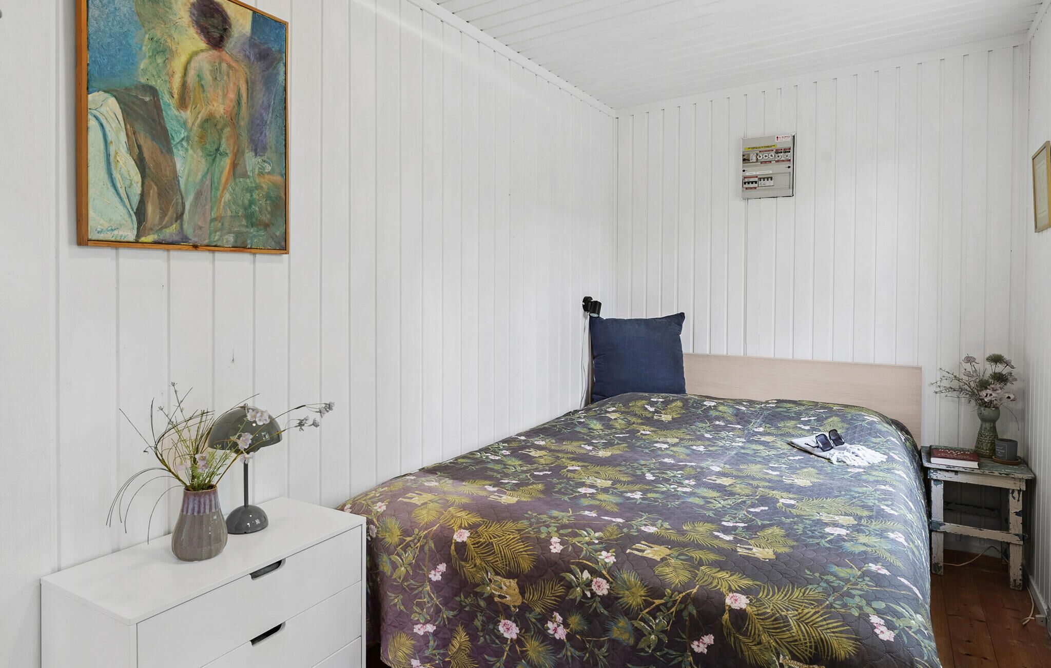 A bedroom with a bed, a nightstand, a dresser, a painting, and a power outlet on the wall.