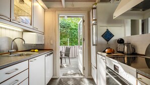Een kleine keuken met een spoelbak, magnetron en oven. Een stoel is te zien buiten de deur, en een raam kijkt uit op groen.