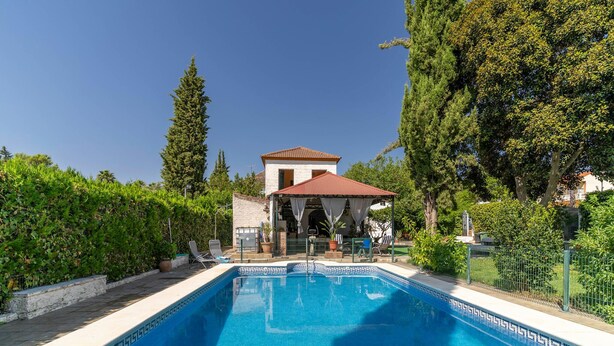 A swimming pool with a covered seating area, surrounded by greenery and a stone building in the background. - Pet friendly home in Guillena (Guillena)