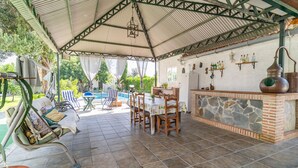 A covered outdoor patio with a stone-tiled floor, a pool, lounge chairs, a dining table, and a bar area with a stone wall and a copper pot. - Pet friendly home in Guillena (Guillena)
