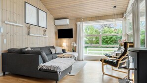 A cozy living room with a grey sofa, a small round table, a wooden floor, a fireplace, and a large window offering a view of greenery. - Nice home in Grevinge with WiFi (Grevinge)