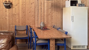 Une pièce aux murs lambrissés, avec une table en bois, des chaises bleues, un canapé en cuir, un réfrigérateur blanc et un luminaire suspendu.