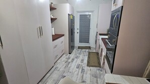 A compact kitchen with white cabinets, a refrigerator, an oven, and a microwave. There is a chair in the foreground and a door leading to another room. - Cozy home away from home apartment. (Bridgetown)