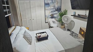 A bedroom with a bed, a desk, a chair, a fan, a painting, and a TV on the wall. - Cozy home away from home apartment. (Bridgetown)