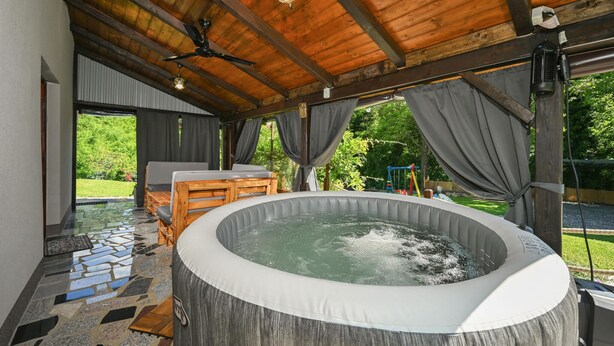 Ein Whirlpool unter einem Holzdach mit Deckenventilator, Fliesenboden und Blick ins Grüne.