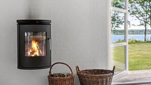 A modern fireplace with a glass front, two wicker baskets, and a view of a lake through a large window. - Amazing home in Roslev with WiFi (Roslev)