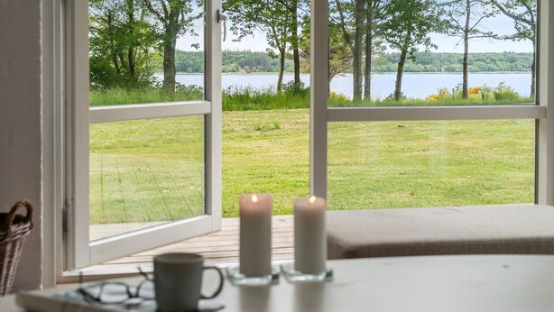 A room with a large window overlooking a grassy area and a body of water. There are two candles on a table in the foreground. - Amazing home in Roslev with WiFi (Roslev)