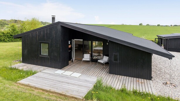 Eine moderne schwarze Holzhütte mit geneigtem Dach, einer Holzdielen-Terrasse und einer Glastür zu einem Innenraum mit zwei Stühlen und einem Tisch.