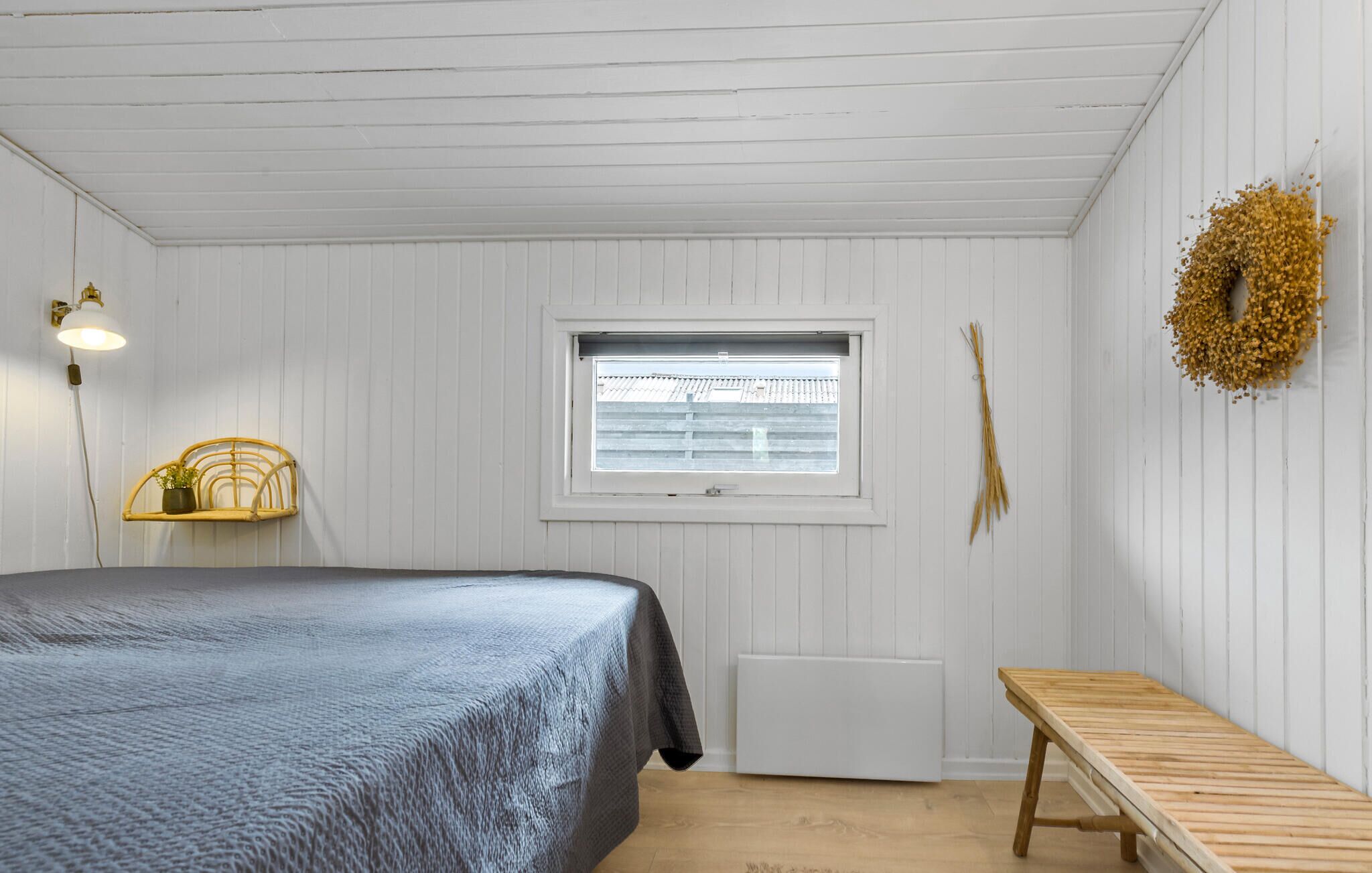 A bedroom with a bed, a bench, a window, a radiator, and a wall-mounted wreath.