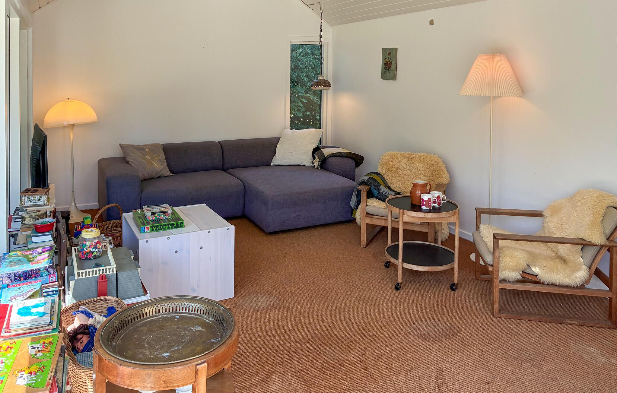 Un salon chaleureux avec un canapé d’angle, une petite table ronde et un fauteuil. On y trouve une lampe, une fenêtre donnant sur un beau paysage et une bibliothèque remplie de divers objets.