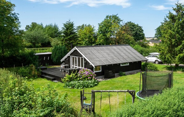 A small, single-story cabin with a sloped roof, surrounded by lush greenery and a fenced yard.