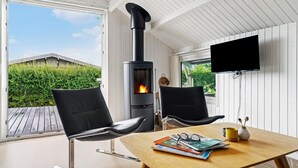 Ein gemütliches Zimmer mit Kamin, zwei schwarzen Stühlen, einem Holztisch mit Zeitschriften, einem an der Wand befestigten Fernseher und Blick ins Grüne.