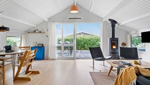 Ein geräumiges Zimmer mit hohen Decken, einem fest eingebauten Holzofen, einer Essecke mit Holzstühlen und einem blauen Schrank sowie einer großen Glasschiebetür, die auf eine Terrassenanlage im Freien führt.